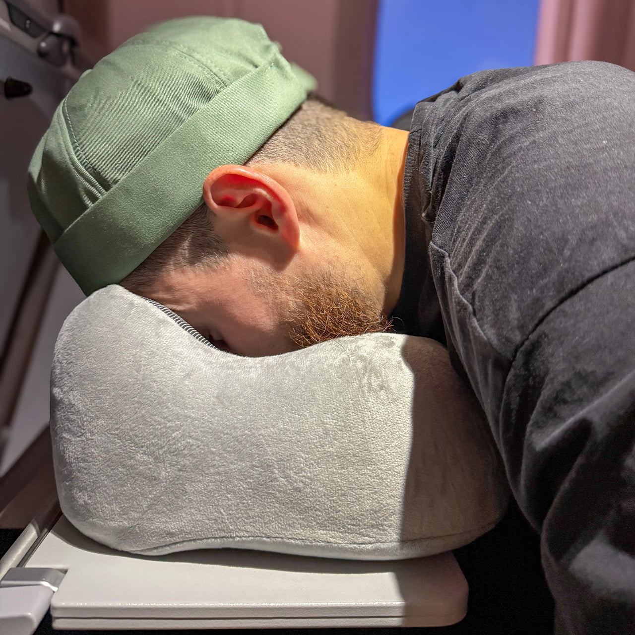 Voyageur utilisant un coussin ergonomique en velours gris perle avec mousse à mémoire de forme en position de repos sur une table d'avion. Idéal pour un confort cervical optimal lors de trajets longs et fatigants.