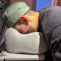 Thumbnail for Voyageur utilisant un coussin ergonomique en velours gris perle avec mousse à mémoire de forme en position de repos sur une table d'avion. Idéal pour un confort cervical optimal lors de trajets longs et fatigants.