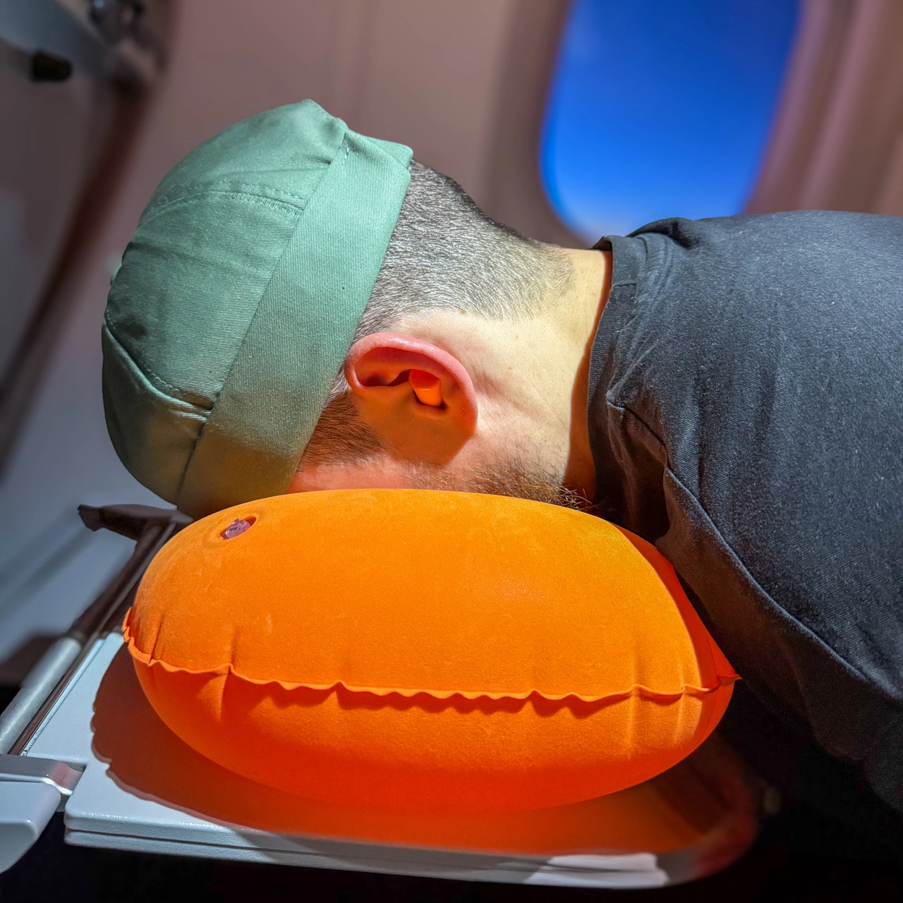 Homme reposant son visage sur un coussin de voyage gonflable orange pendant un vol en avion. Accessoire ergonomique idéal pour un confort et un soutien optimal.