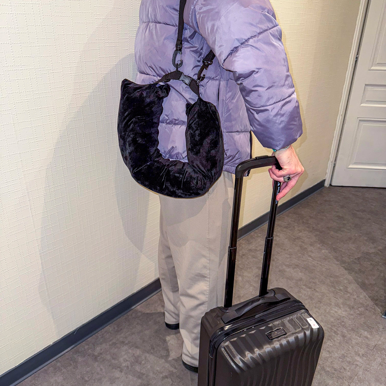 Personne transportant un coussin de voyage noir avec rangement intégré, accroché à l'épaule, tout en tirant une valise. Idéal pour voyager confortablement et léger.