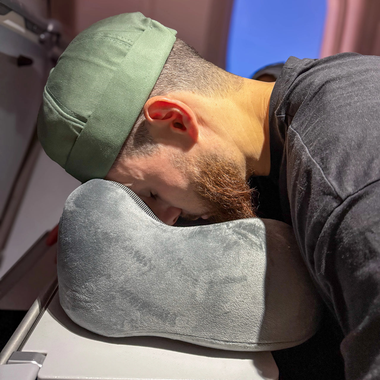 Homme utilisant un coussin de voyage en velours gris anthracite pour dormir confortablement sur une tablette d'avion. Conception ergonomique et velours doux pour un soutien optimal.