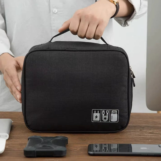Pochette organisateur compacte de couleur noire tenue à la main, posée sur une table en bois avec des accessoires électroniques comme un smartphone et un chargeur, idéale pour le rangement et les voyages.